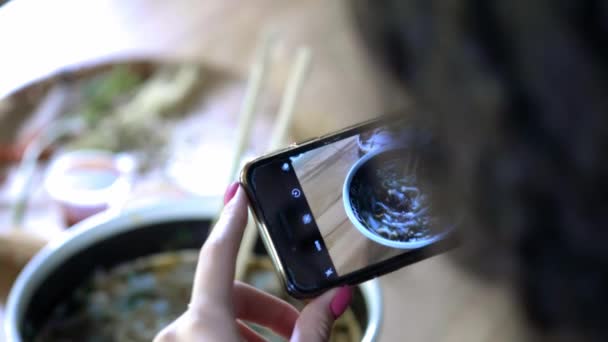 Ein Mädchen macht ein Foto von Lebensmitteln auf dem Smartphone. Teilen Sie mit Freunden ein Foto von Essen in sozialen Netzwerken. Café mit asiatischem Essen. sozial aktiver Lebensstil, virtuelle Kommunikation mit Freunden. — Stockvideo