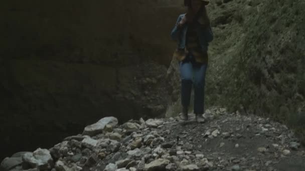 Fille voyageuse dans une grotte ou un canyon. Elle porte un chapeau et une veste et a un sac à dos. Explore une ancienne grotte, à la recherche d'aventure. Randonnée pédestre à travers une étroite allée dans une montagne . — Video
