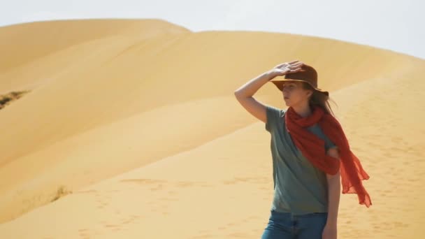 Retrato: Chica viajera en el desierto sobre la arena. Llevando un sombrero y una bufanda roja. Explora terrenos antiguos, en busca de aventura. El viento sopla. Estilo de vida activo, tiene una pasión por la aventura . — Vídeo de stock