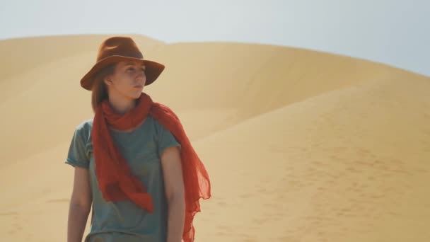Portret: meisje reiziger in de woestijn op het zand. Het dragen van een hoed en een rode sjaal. Verkent het oude terrein, op zoek naar avontuur. De wind waait. Actieve levensstijl, heeft een passie voor avontuur. — Stockvideo