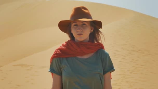 Retrato: Chica viajera en el desierto sobre la arena. Llevando un sombrero y una bufanda roja. Explora terrenos antiguos, en busca de aventura. El viento sopla. Estilo de vida activo, tiene una pasión por la aventura . — Vídeo de stock