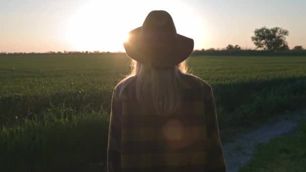 Uma jovem encantadora viajante ou agricultor olha para o pôr do sol ou nascer do sol. Vestindo camisa xadrez e chapéu. Mulher despreocupada bonita desfrutando da natureza e da luz solar no campo de trigo no incrível pôr do sol colorido . — Vídeo de Stock