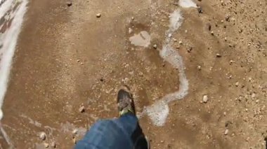 Adam yavaş hareket kumlu plajda çıplak ayak yürüyüş. Bir adam sahil boyunca sahil boyunca yürür. Yaz ayakkabıları giyiyor. Tuzlu suda ıslatılmış ayakkabılar. Denizde Yaz yürüyüşü.