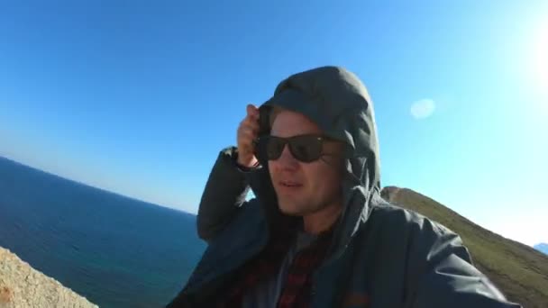 フードの若いスタイリッシュな男。岬に立っている。海の周り。旅行とアクティブなライフスタイルを楽しむ. — ストック動画