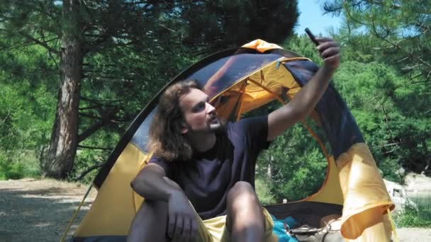 Un hombre está sentado en una tienda de campaña en un bosque verde cerca del lago. Realiza una videollamada en un smartphone a amigos o novia. Le muestra un hermoso lugar junto al lago con una tienda de campaña . — Vídeos de Stock