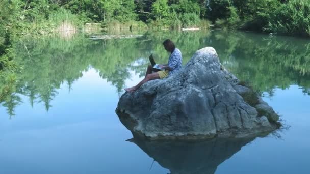 Un joven freelance se sienta en una piedra en medio de un lago con un portátil. Concepto independiente. Trabajo en viajes. Internet inalámbrico en el bosque — Vídeos de Stock