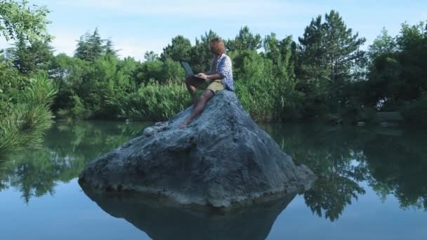 Een jonge freelance man zit op een steen in het midden van een meer met een laptop. Freelance concept. Werken aan reizen. Draadloos internet in het bos — Stockvideo