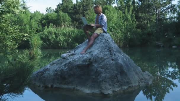 젊은 프리랜서 남자가 노트북을 들고 호수 한가운데에 있는 돌 위에 앉아 있다. 프리랜서 개념입니다. 여행 에 대한 작업. 숲속의 무선 인터넷 — 비디오