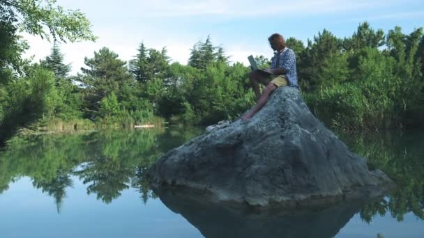 Egy fiatal szabadúszó srác ül egy kő közepén egy tó egy laptoppal. Szabadúszó koncepció. Munka az utazással. Vezeték nélküli internet az erdőben — Stock videók