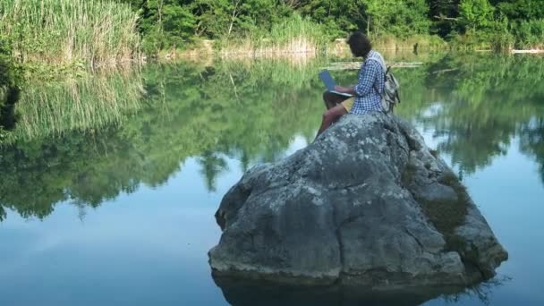 一个年轻的自由职业者坐在湖边的一块石头上，拿着一台笔记本电脑。自由职业者的概念。工作旅行。森林中的无线互联网 — 图库视频影像