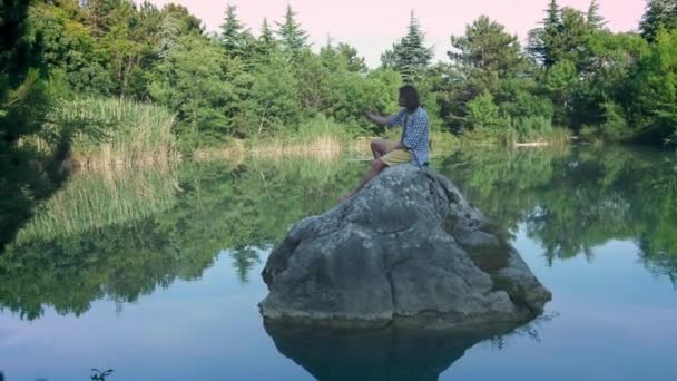 A young guy sits on a stone in the middle of the lake, takes a selfie. Uses a smartphone for photography. The concept of travel. Wireless internet in the forest — Stock Video