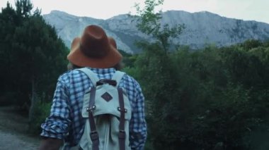 Dağların arka planında ormanda yürüyüş eden genç bir adam. Bir akıllı telefonda dağların bir fotoğrafını yapar. Aktif yaşam tarzı. Damalı gömlek ve gezgin şapkası giyiyor. Yürüyüş seyahat konsepti