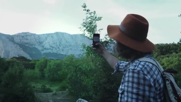 산을 배경으로 숲에서 하이킹하는 젊은 남자. 스마트폰으로 산사진을 만듭니다. 활동적인 라이프 스타일. 체크 셔츠와 여행자 모자를 착용. 하이킹 여행 컨셉 — 비디오