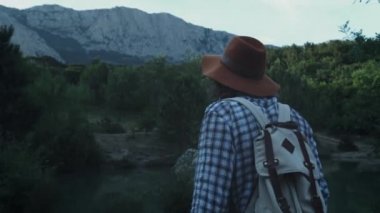 Dağların arka planında ormanda yürüyüş eden genç bir adam. Aktif yaşam tarzı. Damalı gömlek ve gezgin şapkası giyiyor. Yürüyüş seyahat konsepti