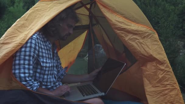 Homme indépendant travaillant avec un ordinateur portable assis dans une tente de camping sur la forêt. Freelance travaillant sur un nouveau projet de démarrage en utilisant un ordinateur portable et une connexion sans fil. Voyages d'été freelance . — Video