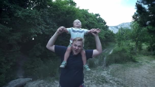 Happy young father holds his son piggyback ride on his shoulders and looking up. Little boy is sitting piggyback on shoulders his dad while imitating the flight. — Stock Video