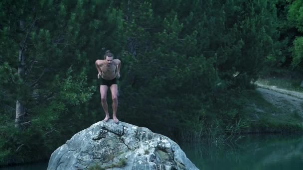 En ung kille på en sommarsemester eller semester hoppar från en sten i mitten av en fjäll skog sjö. Hoppar in i det kalla fräscha klara vattnet, en naturlig gatulivsstil. — Stockvideo