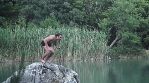 Ein junger Kerl im Sommerurlaub oder im Urlaub, der von einem Stein inmitten eines Bergwaldsees springt. Sprünge ins kalte, klare Wasser, ein natürlicher Lebensstil auf der Straße. — Stockvideo