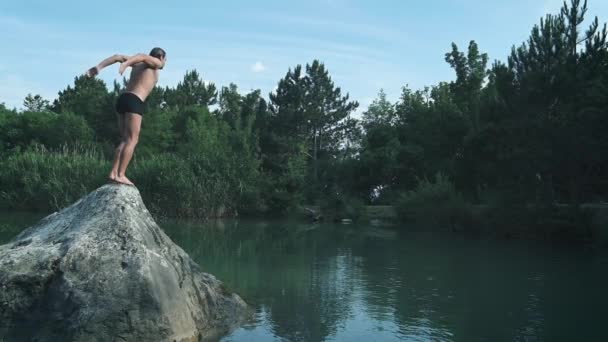 Um cara jovem em umas férias de verão ou salto de férias de uma pedra no meio de um lago de floresta montesa. Salta para a água fria e fresca, um estilo de vida natural de rua . — Vídeo de Stock
