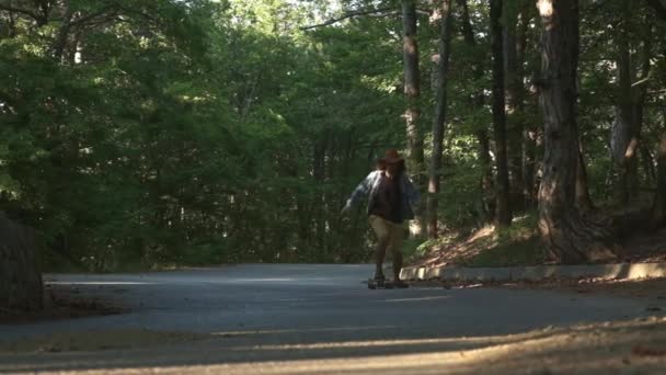 En ung kille lär sig att rida en longboard. Det ligger i skogen. Klädd i en pläd skjorta och en filt hatt. Aktiv livsstil. — Stockvideo