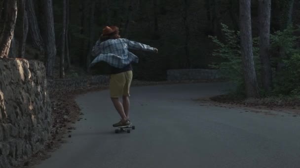 Młody facet uczy się jeździć na longboard. Znajduje się on w lesie. Ubrany w koszulę i kapelusz z filcu. Aktywny styl życia. — Wideo stockowe