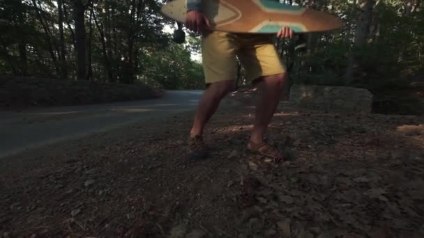 Nuori hipsteri metsässä pilkkaamassa longboardin kanssa. Käyttää longboardia kitarana. Hyppää kuin rokkitähti. Hauskaa ajanvietettä kesälomilla. Hidastettu laukaus . — kuvapankkivideo