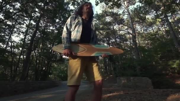 Jeune hipster dans la forêt, se moquant d'un longboard. Utilise le longboard comme guitare. Il saute comme une rock star. passe-temps amusant pendant les vacances d'été. Plan au ralenti . — Video