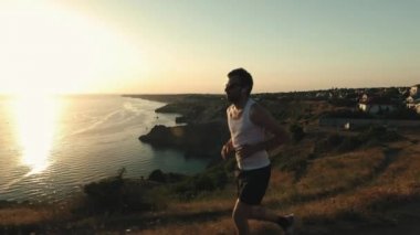 Athletic Handsome Man Telefon ve Kulaklık ile Spor yapıyor, Yaz plajda Jogging sırasında Günbatımında Sea Coast boyunca Çalışan. Kablosuz kulaklık kullanır.
