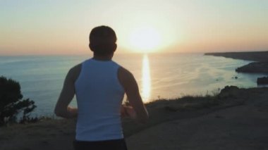 Athletic Handsome Man Telefon ve Kulaklık ile Spor yapıyor, Yaz plajda Jogging sırasında Günbatımında Sea Coast boyunca Çalışan. Kablosuz kulaklık kullanır.