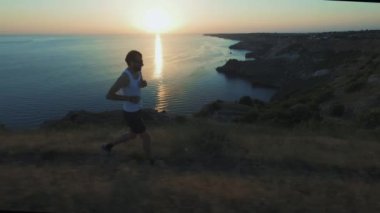 Athletic Handsome Man Telefon ve Kulaklık ile Spor yapıyor, Yaz plajda Jogging sırasında Günbatımında Sea Coast boyunca Çalışan. Kablosuz kulaklık kullanır.
