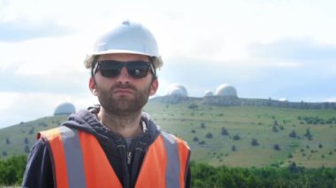 Erkek mühendis veya beyaz bir kask ve yelek içinde oluşturucu gözlük giyer. Yetiştiricilerin birkaç yarımkürenin arka planında. Yıldızları ve havayı izlemek.