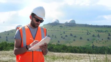 Beyaz kasklı ve yelekli bir erkek mühendis belgeleri doldurur, elinde kağıt kağıtları tutar. Yetiştiricilerin birkaç yarımkürenin arka planında. Yıldızları ve havayı izlemek.
