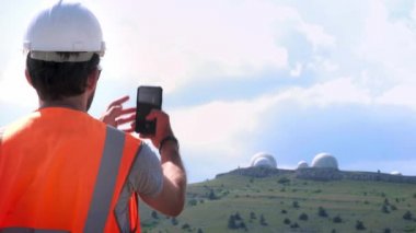 Beyaz kask ve yelekli erkek bir mühendis veya inşaatçı dağın tepesindeki gözlemevinin fotoğrafını çeker. Planitaria'nın birkaç yarımküresi. Yıldızları ve havayı izlemek