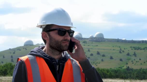 Male engineer or builder in a white helmet and talking on the phone, making a call. Against the backdrop of several hemispheres of the planters. Watching the stars and the weather — Stock Video