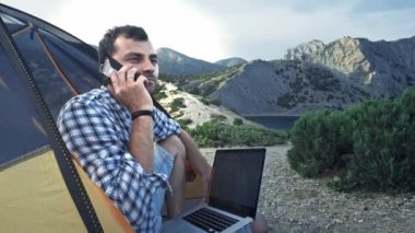 Sahilde Bir Kamp Çadırında Oturan Laptop Kullanarak Çalışan Freelancer Man. Telefonda konuşuyorum. Freelancer kablosuz bağlantı kullanarak yeni başlangıç projesi üzerinde çalışıyor. Serbest Yaz Seyahat.