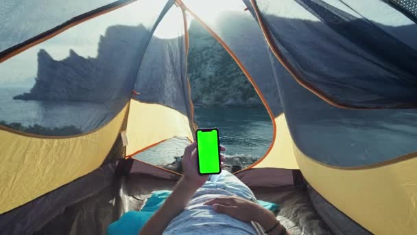 Le panneau solaire fixé à la tente. L'homme assis à côté des frais de téléphone portable du soleil. Un jeune homme se trouve dans une tente de camp, tenant un smartphone avec un écran vert. Camp tente se dresse sur la plage . — Video