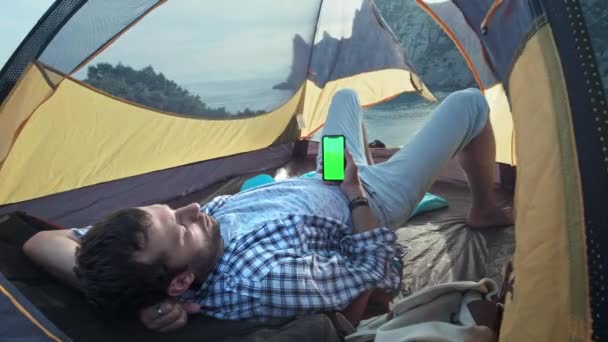 El panel solar unido a la carpa. El hombre sentado al lado del teléfono móvil carga desde el sol. Un joven yace en una tienda de campaña, sosteniendo un teléfono inteligente con una pantalla verde. Tienda de campaña en la playa . — Vídeos de Stock