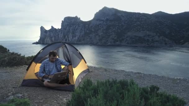 Uomo freelance che lavora con il computer portatile seduto in una tenda da campeggio sulla spiaggia. Freelancer che lavora su un nuovo progetto di startup utilizzando computer portatile e connessione wireless. Viaggi estivi freelance — Video Stock