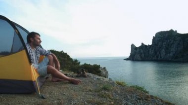 Genç bir adam kamp çadırında oturuyor. Akıllı telefonda fotoğraf yapar. Deniz ve dağların güzel manzara bir çekim yapar. Trekking gününün ardından plajda dinlendirici nin keyfini çıkarabilirsiniz. Yürüyüş gezisi