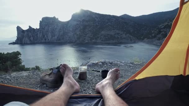 En ung kille ligger i ett läger tält, benen sticker ut från tältet. Njuter av att koppla av på stranden efter vandrings dagens äventyr. Läger tält står på stranden. Vandringstur i Krim. — Stockvideo