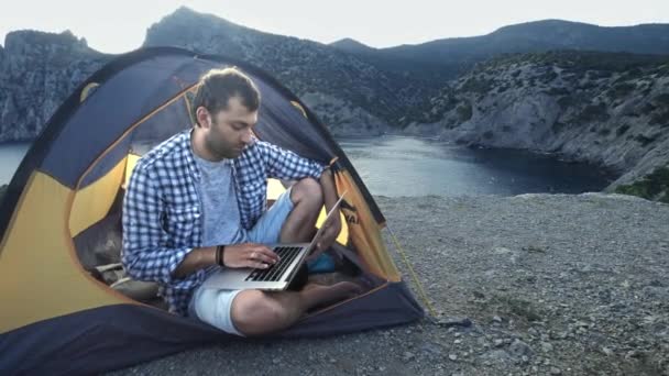 Freelancer Man Working Using Laptop Sitting In A Camping Tent On The Beach. Freelancer que trabaja en un nuevo proyecto de inicio con ordenador portátil y conexión inalámbrica. Viajes de verano independientes . — Vídeos de Stock