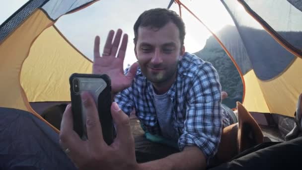 Um homem faz uma chamada de vídeo para amigos, parentes ou namorado para mostrar-lhe o belo lugar onde acampar com a tenda. Tenda de acampamento fica na praia . — Vídeo de Stock