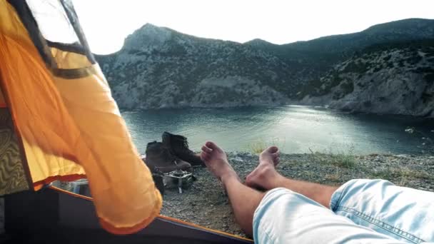 Genç bir adam bir kamp çadırında yatıyor, bacakları çadırdan çıkıntı. Trekking gününün macerasının ardından sahilde dinlenmenin keyfini çıkarabilirsiniz. Kamp çadırı deniz kıyısında duruyor. Kırım'da yürüyüş gezisi. — Stok video