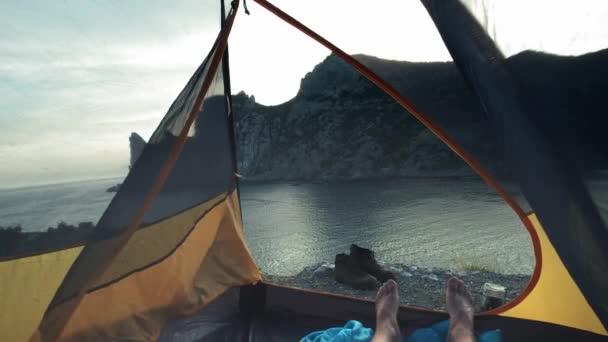 一个年轻人躺在一个营地的帐篷里，他的双腿从帐篷里伸出来。徒步旅行后，您可以在海滩上放松身心。营地帐篷矗立在海边。克里米亚徒步旅行. — 图库视频影像