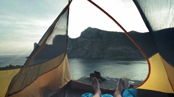 若い男がキャンプテントに横たにあり、彼の足はテントから突き出ています。トレッキングデーの冒険の後はビーチでリラックスできます。キャンプテントは海岸に立っています。クリミアでのハイキング旅行. — ストック動画