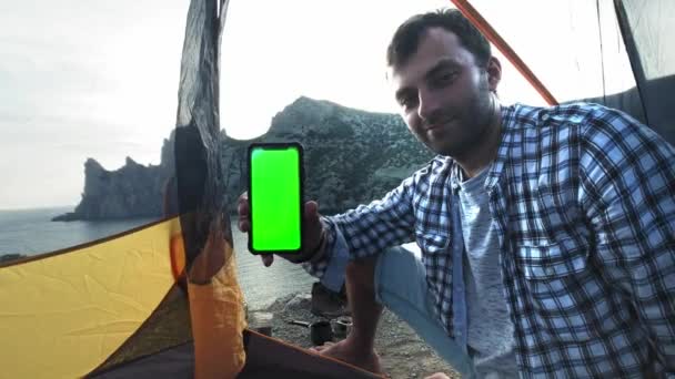 Le panneau solaire fixé à la tente. L'homme assis à côté des frais de téléphone portable du soleil. Un jeune homme se trouve dans une tente de camp, tenant un smartphone avec un écran vert. Camp tente se dresse sur la plage . — Video