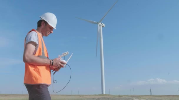 En man styr en drönare i ett fält på bakgrunden av ett vindkraftverk. Inspekterar vindkraftverket med en copter. Begreppet miljöförorening, ny teknik för alternativ energi — Stockvideo