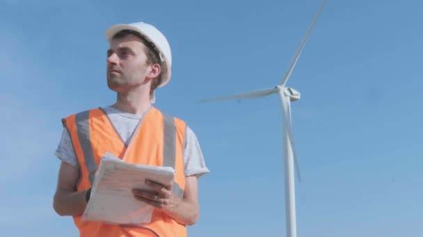 Um engenheiro masculino preenche documentos em um campo em meio a uma turbina eólica. Inspeciona a turbina eólica com um helicóptero. O conceito de poluição ambiental, novas tecnologias de energia alternativa — Vídeo de Stock