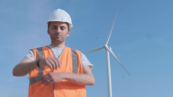 PORTRAIT: Close shot of adult engineer in orange vest and white hard hat with wind turbines on background looking at camera. — Stock Video