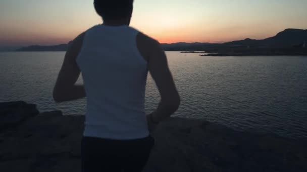 Attractive young man running athlete runner training tough cardio exercise workout on calm ocean seaside run wearing earphones. Healthcare, active lifestyle, sunset dusk and twilight. — Stock Video